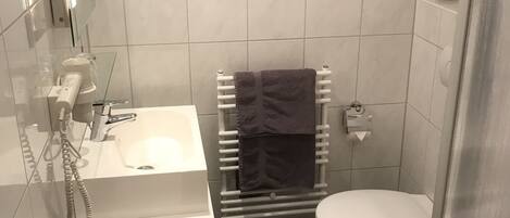 A compact bathroom with a glass-enclosed shower, a white sink with a handheld showerhead, a towel rack with a purple towel, a toilet, and a small window above the sink.