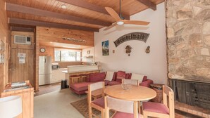 A cozy cabin interior with a wooden ceiling, a stone wall, a dining area with a round table, and a kitchen with a refrigerator and microwave.
