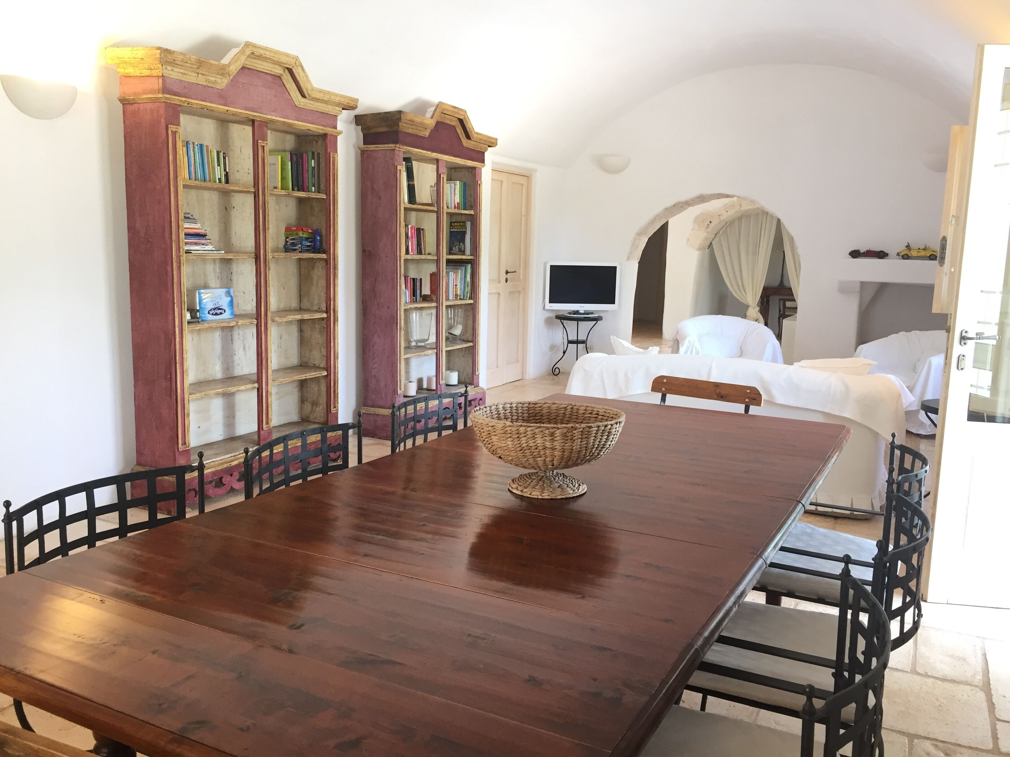 A dining area with a wooden table and chairs, two bookshelves with books, a television, and a white sofa.