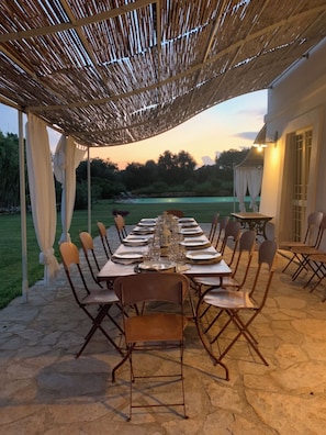 An outdoor dining area with a long table set for a meal, surrounded by chairs, under a thatched roof structure.