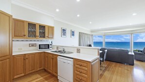 Cozinha moderna com armários de madeira, micro-ondas e lava-louças. Há uma área de jantar com sofá cinzento e vista para o oceano através de grandes janelas.