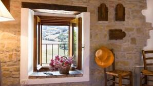 Quarto rústico com parede de pedra, janela com vista para colinas ondulantes, cesta de vime com flores e duas cadeiras de madeira.