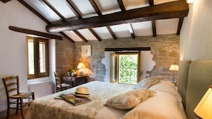 Quarto com parede de pedra, vigas de madeira, cama com almofadas, uma cadeira, escrivaninha com abajur e uma janela.