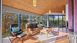 A spacious living area with a leather sofa, a black leather chair, and a wooden coffee table. Large windows offer a view of the outdoors. - 6 Acacia St Sandy Point (Sandy Point)