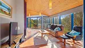 A spacious living room with a large TV, comfortable seating, and a view of the outdoors through large windows. - 6 Acacia St Sandy Point (Sandy Point)