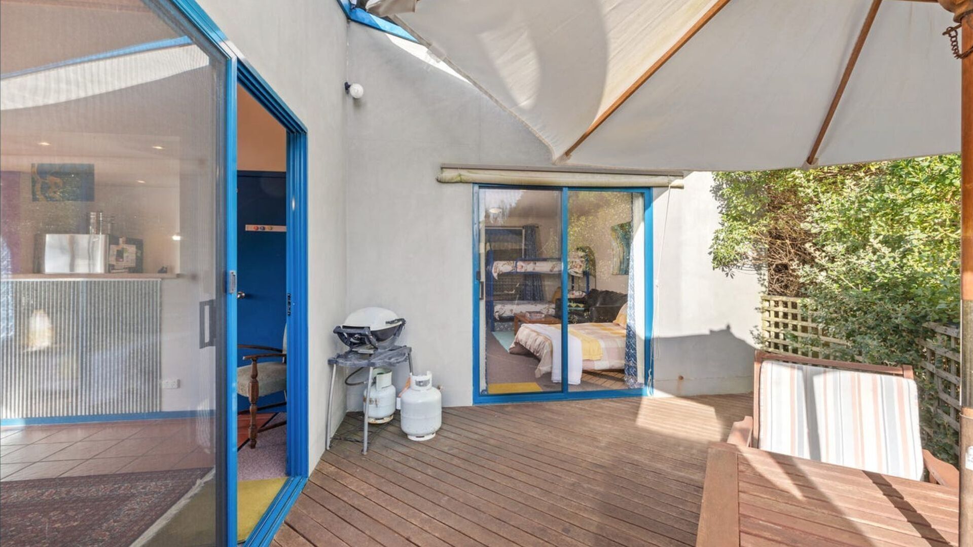 Un espace extérieur moderne doté d’une terrasse en bois, d’un coin salon couvert et d’une porte vitrée donnant accès à une chambre avec un lit et un bureau.