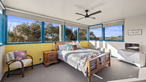 A bedroom with a bed, a chair, a nightstand, a TV, and large windows with a view of trees. - 6 Acacia St Sandy Point (Sandy Point)