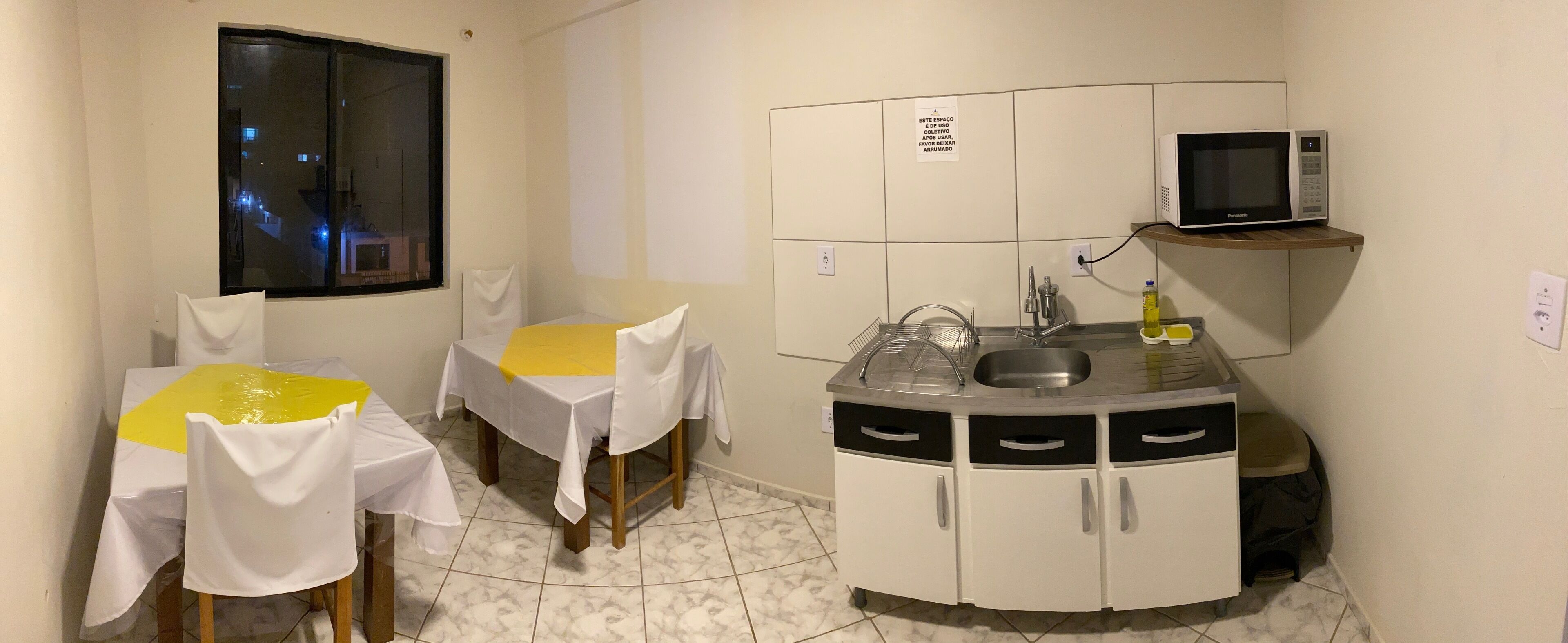 Cocina compacta con microondas, lavabo y armarios de almacenamiento. Hay dos mesas de comedor con sillas, y una ventana con vistas al exterior.