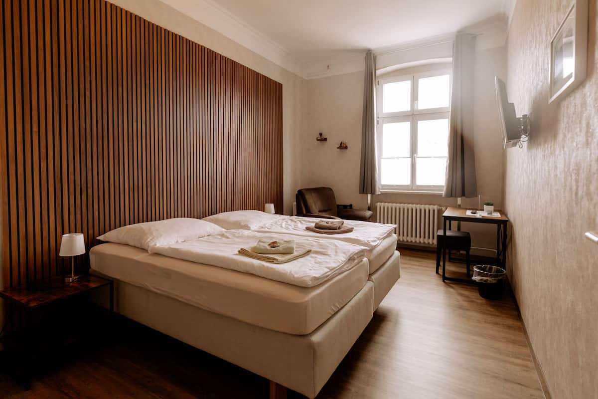 A hotel room with a large bed, a desk, a chair, a radiator, and a window with curtains.