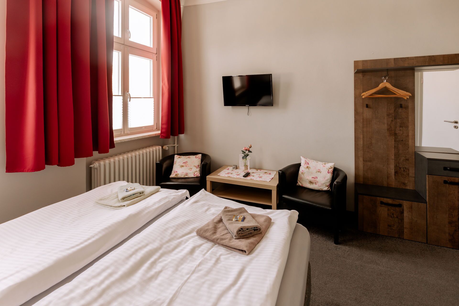 A hotel room with two beds, a TV, a wooden wardrobe, a small table with a vase, and a red curtain.