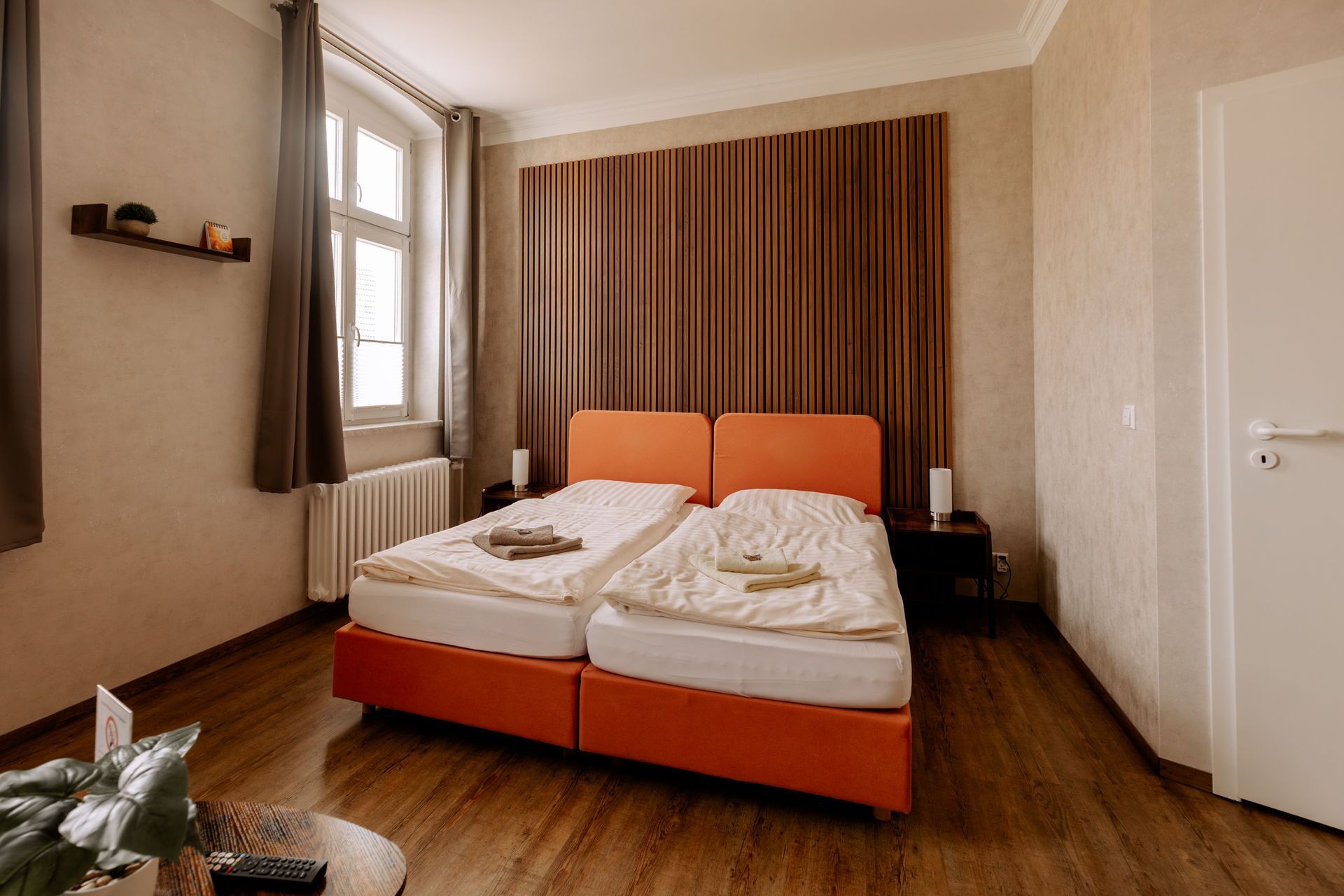 A double bed with an orange headboard, a bedside table with a lamp, a window with curtains, and a wooden slatted wall.