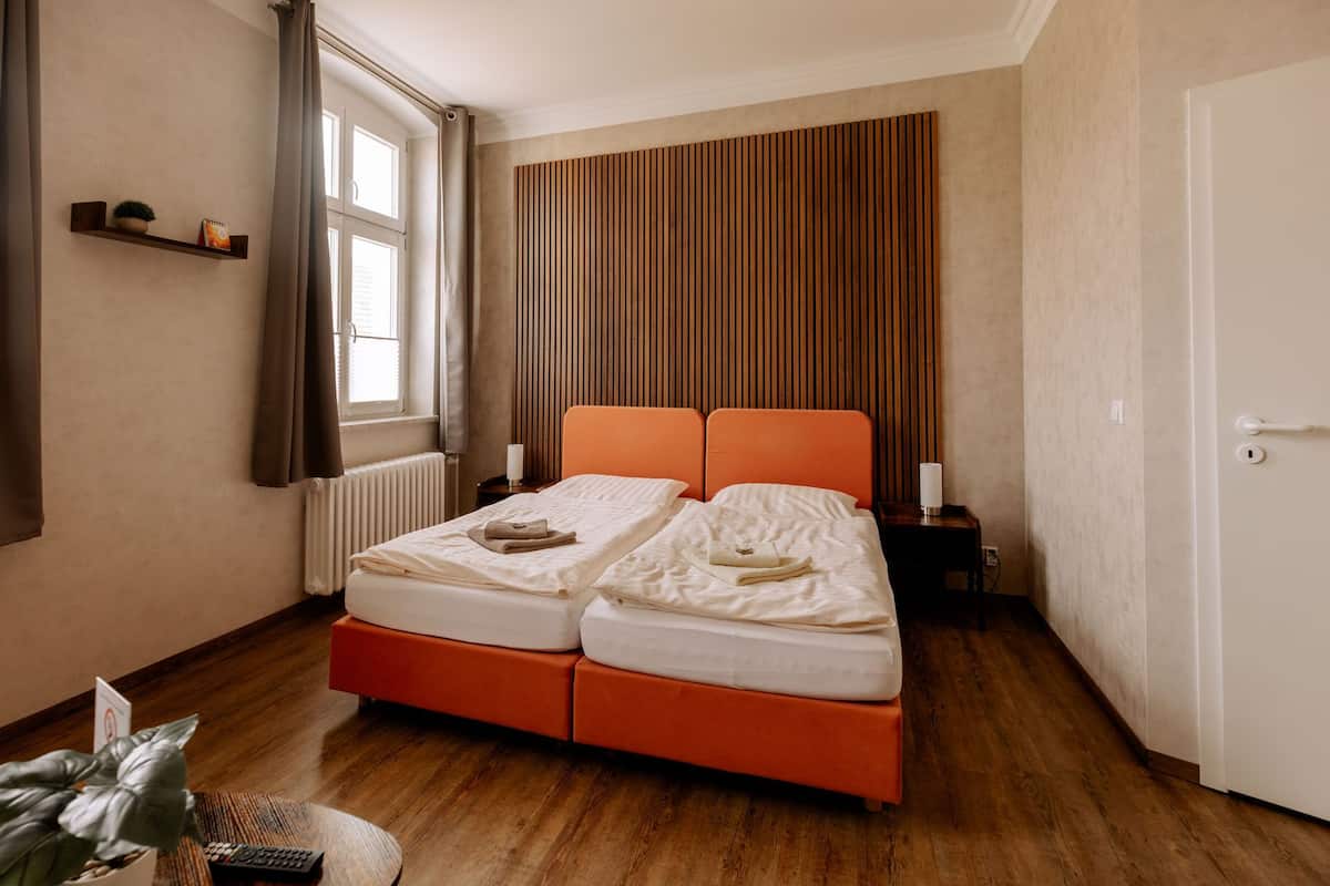 A double bed with an orange headboard, a bedside table with a lamp, a window with curtains, and a wooden slatted wall.