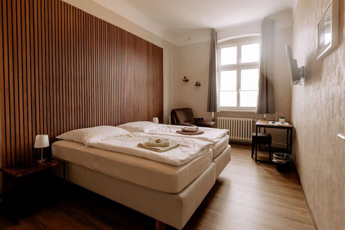 A hotel room with a bed, a desk, a chair, a window, and a radiator.