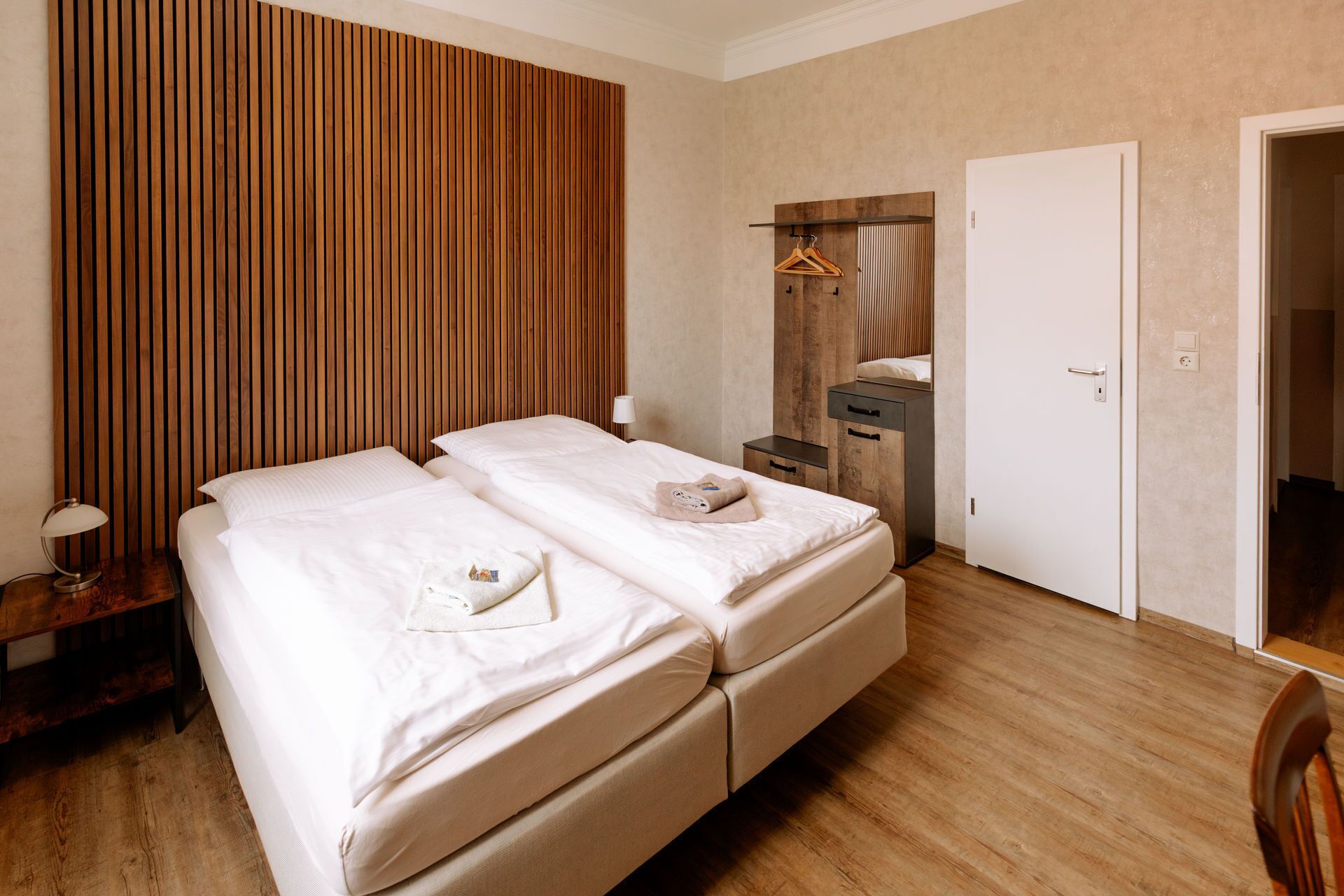 Habitación de hotel con pared de listones de madera, cama con ropa blanca, mesita de noche con lámpara, cómoda y una puerta.