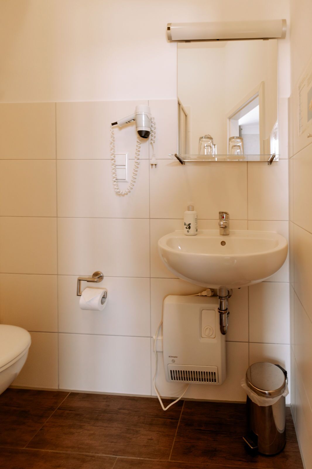 Un baño con lavamanos blanco, espejo, secador de pelo, inodoro y cubo de basura.