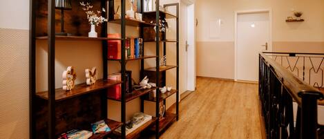 A hallway with wooden flooring, a staircase with a black railing, and a bookshelf with various items including books and decorative objects.