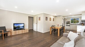 Ett modernt vardagsrum med en platt-TV, möbler i trä, en matplats med bord och stolar samt ett kök med vitvaror i rostfritt stål.