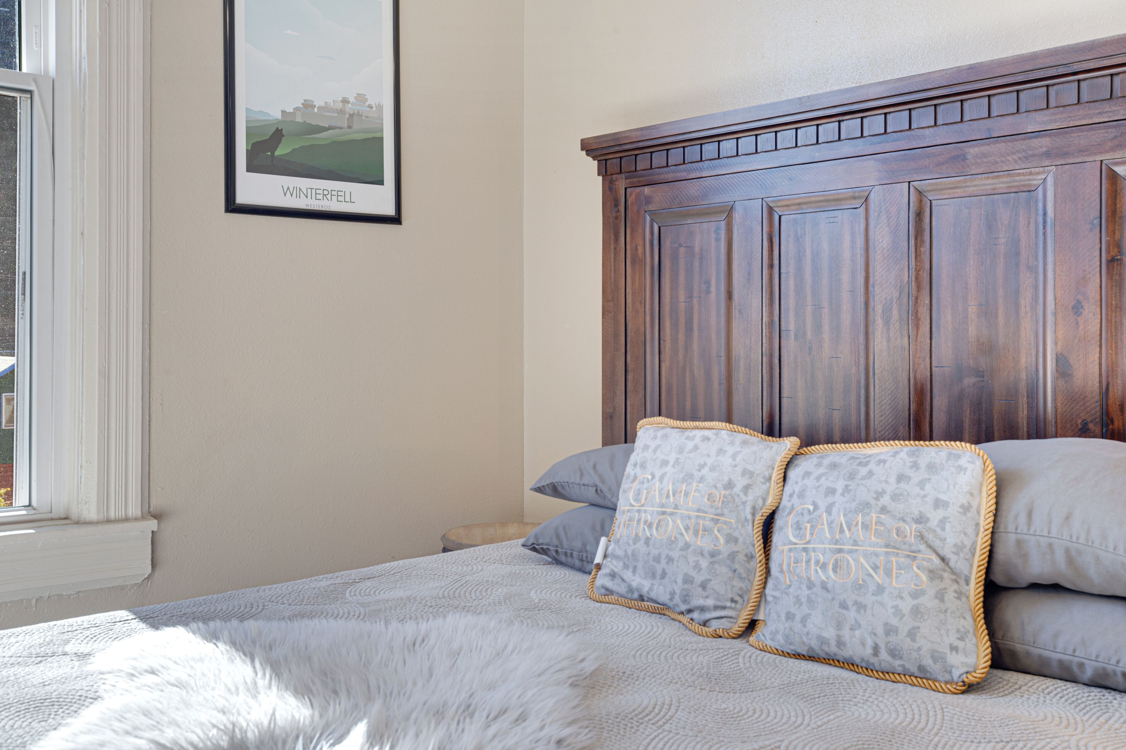 Een slaapkamer met een groot houten hoofdeinde, een lijst met foto aan de muur en twee kussens met een Game of Thrones dessin.