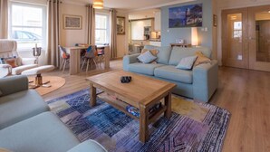 A modern living room with a sofa, a coffee table, and a rug. There is a dining area with chairs and a table. A kitchen is visible in the background. - Marina View Apartment (Inverkip)
