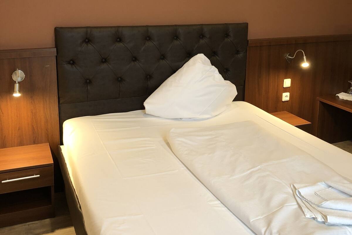 Un lit bien fait, avec du linge de lit blanc et une tête de lit capitonnée de couleur sombre, dans une chambre d’hôtel.