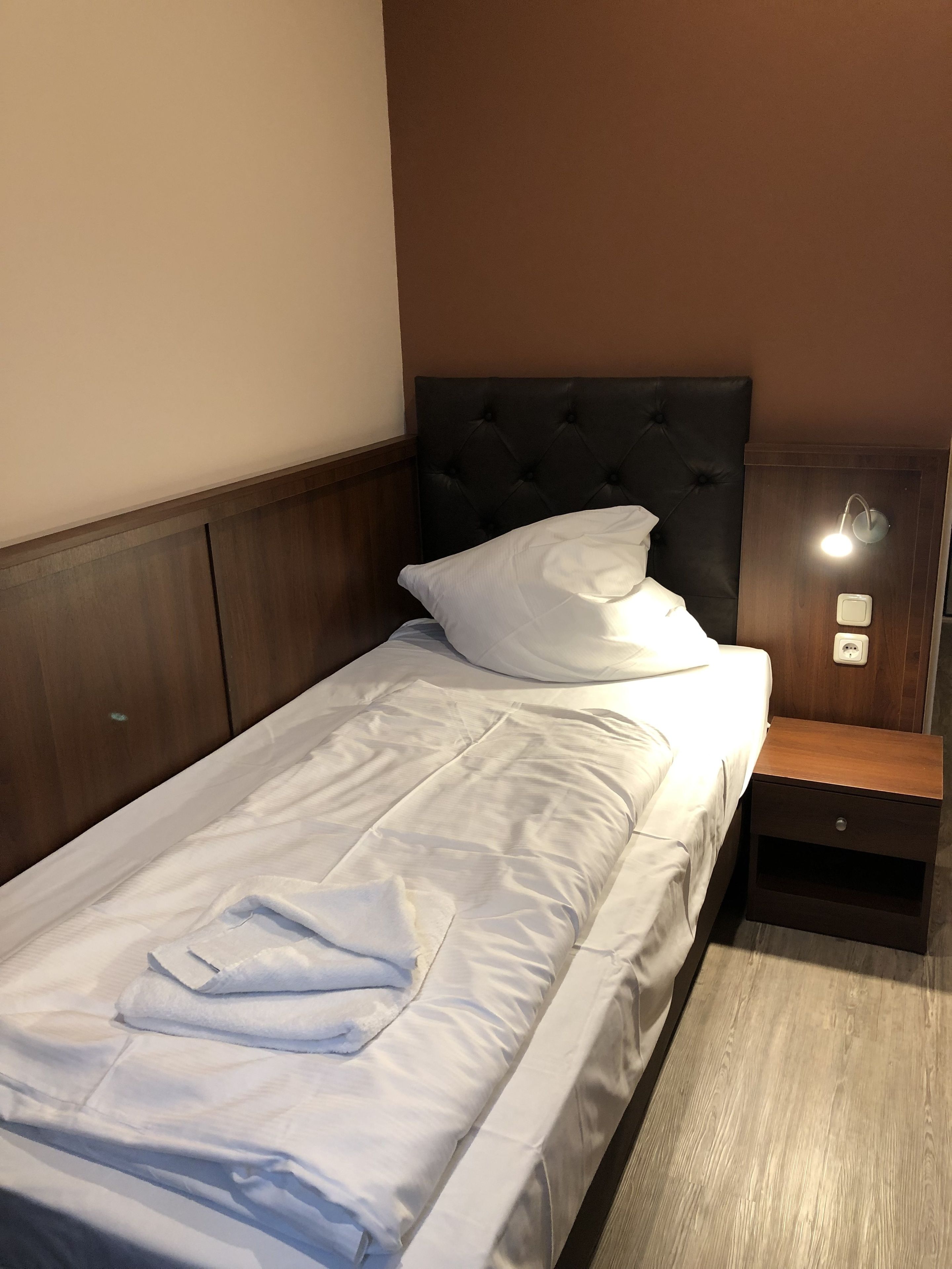 A single bed with white linens, a wooden headboard, a nightstand with a lamp, and a wall socket.