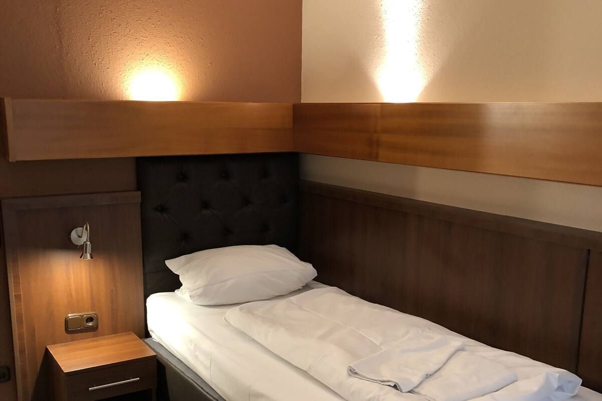 Un lit simple avec une tête de lit en bois, une table de chevet avec une lampe et une petite commode.