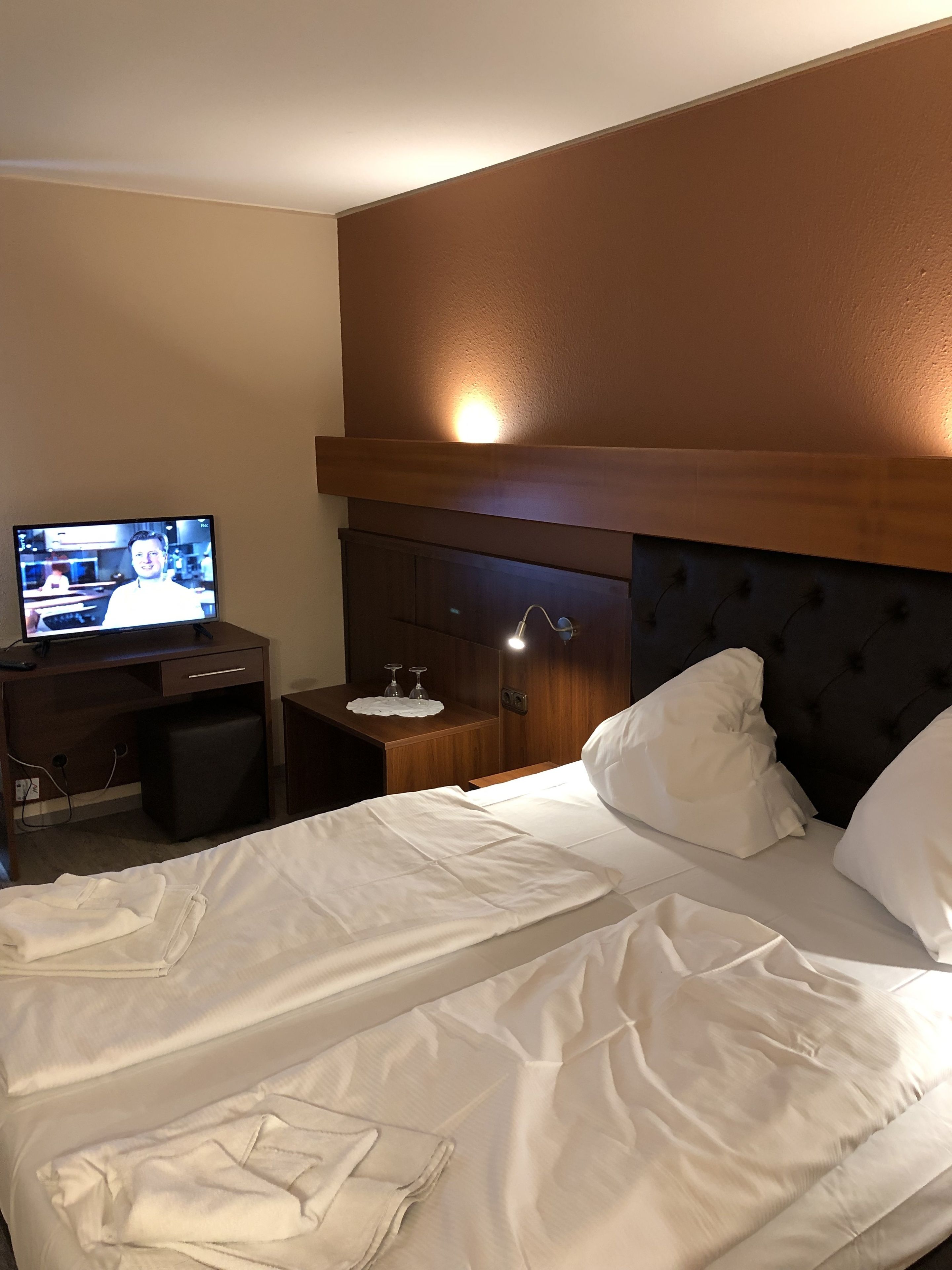 A hotel room with a bed, a desk with a television, a nightstand, and a headboard.
