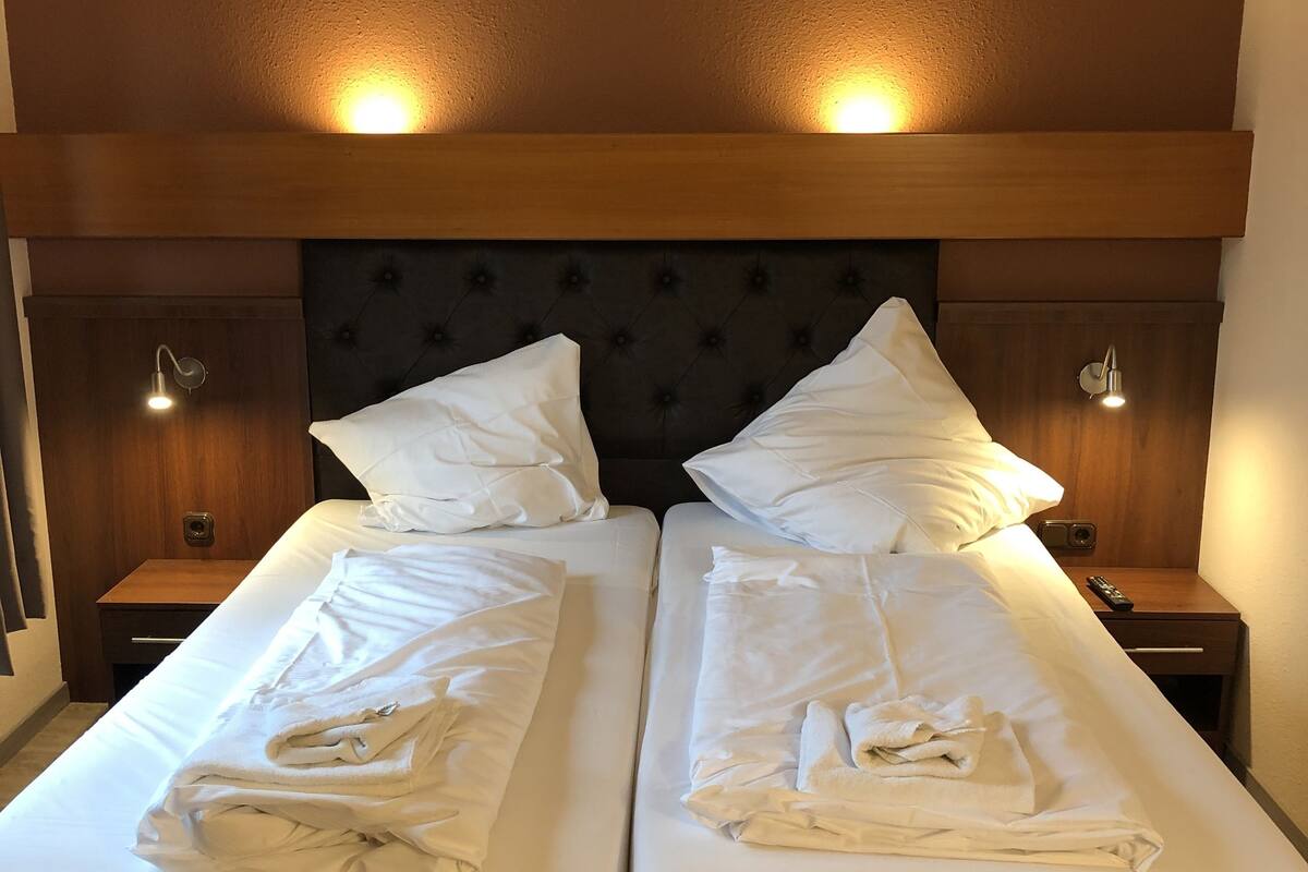 Un lit double avec du linge de lit blanc et une tête de lit en bois. Il y a deux tables de chevet avec des lampes, et la pièce est décorée dans des tons chauds de brun.