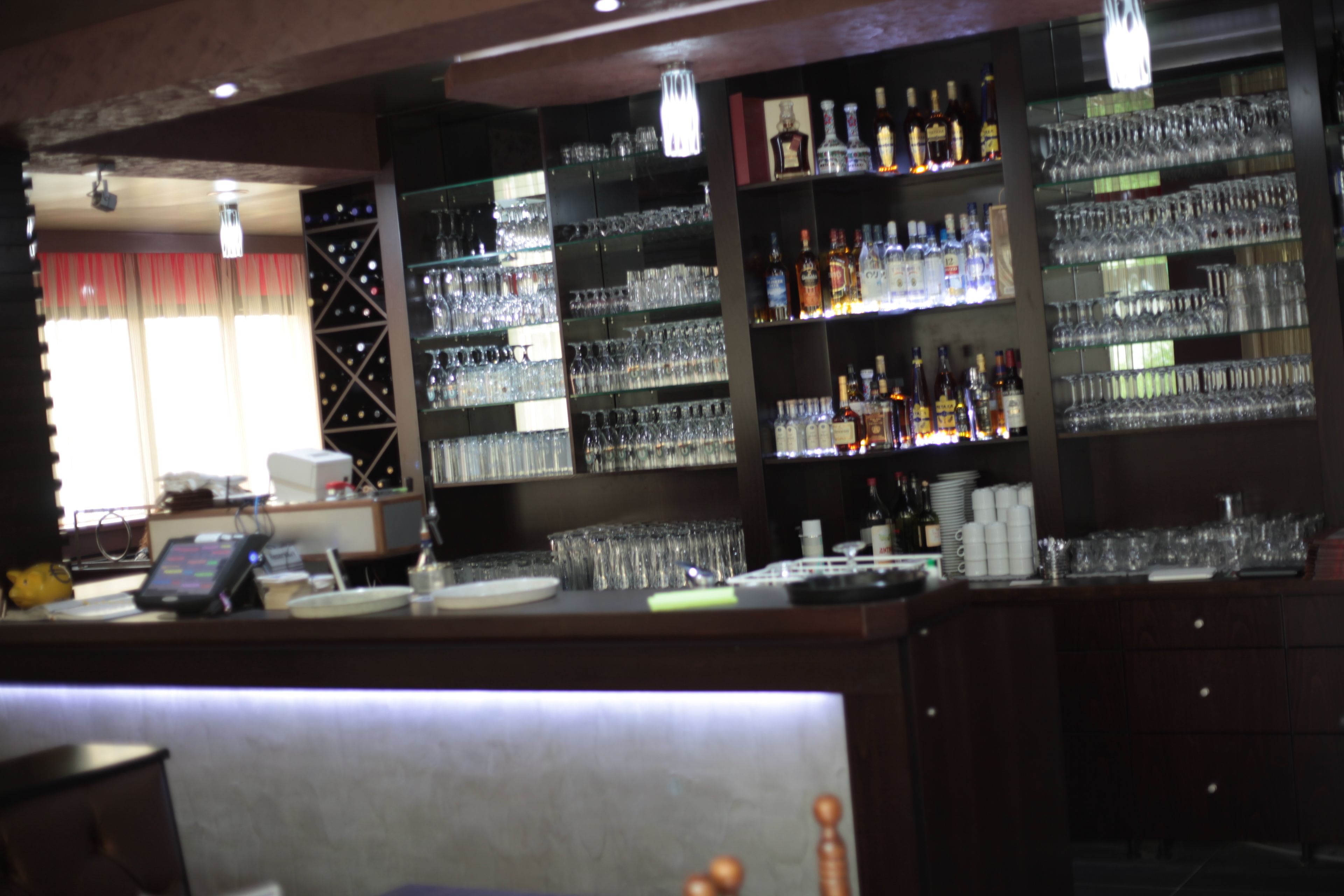 A bar area with shelves of bottles and glasses, a cash register, and a counter.
