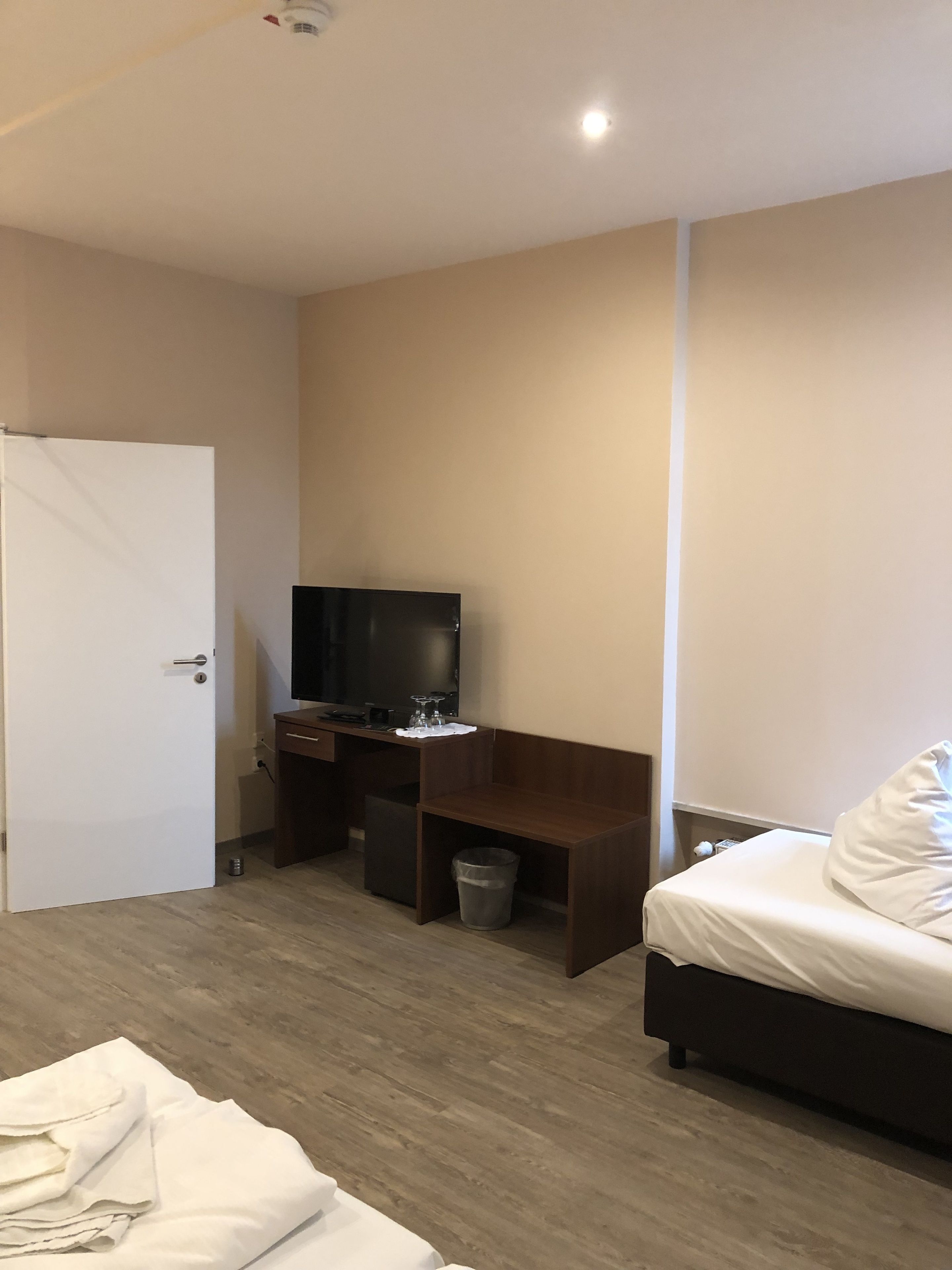 A bedroom with a bed, a desk with a TV, a trash bin, and a door.