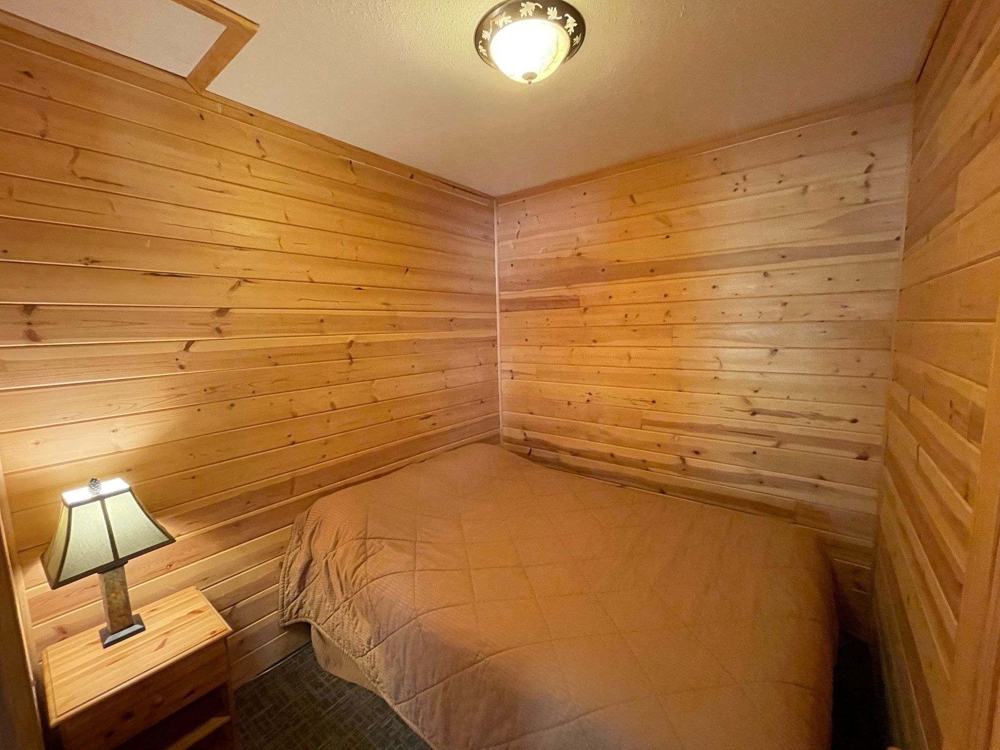 Una habitación pequeña y acogedora con paredes de madera y una cama individual. Hay una mesita de noche con una lámpara y una pequeña mesa.