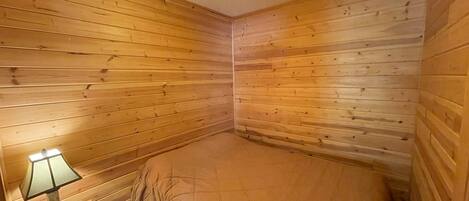 Una habitación pequeña y acogedora con paredes de madera y una cama individual. Hay una mesita de noche con una lámpara y una pequeña mesa.