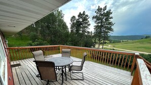 A wooden deck with a round table and four chairs, overlooking a green landscape with trees and a golf course.