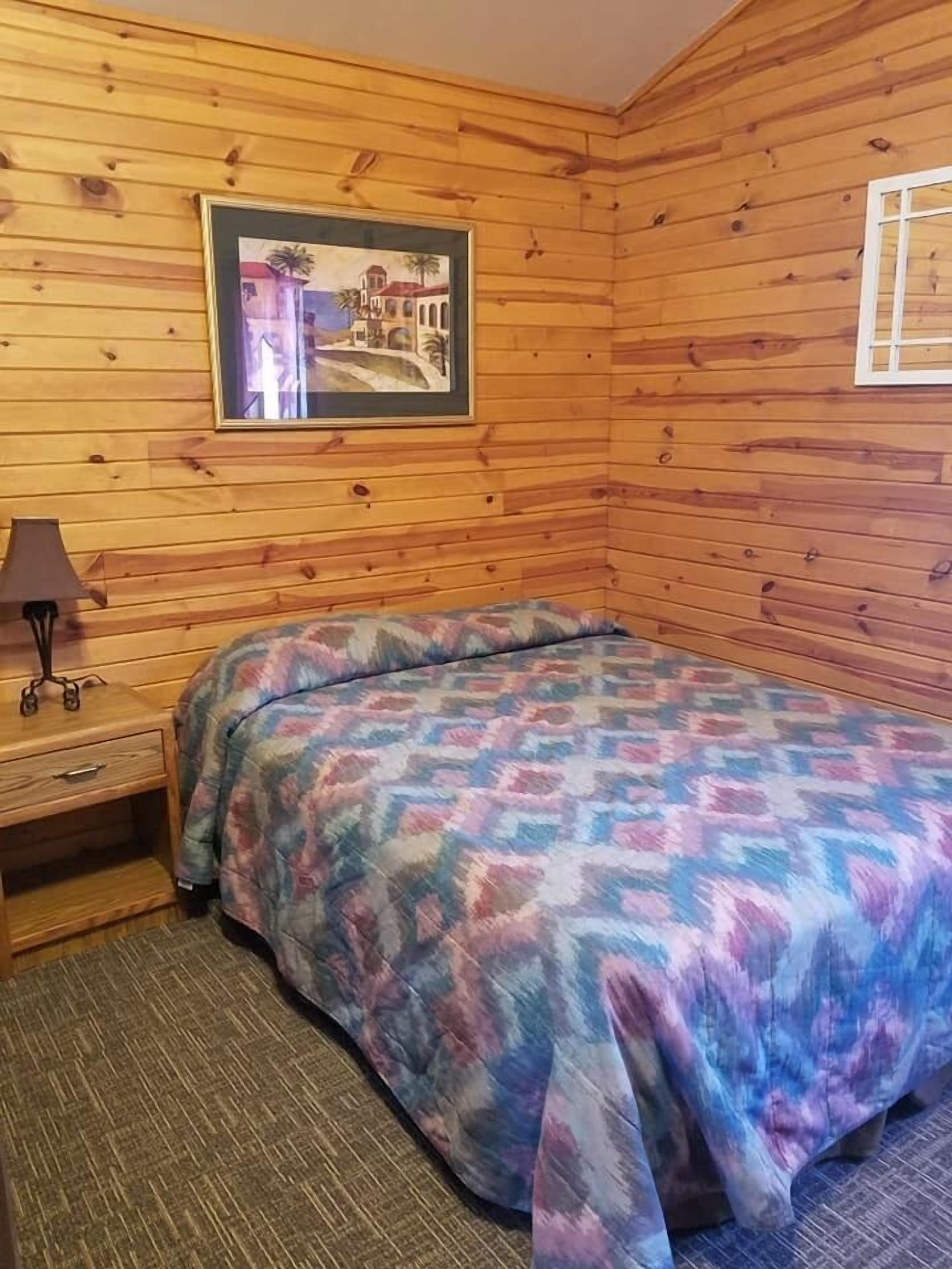 Una habitación con piso de madera, una cama, una mesita de noche, una lámpara y un cuadro en la pared.