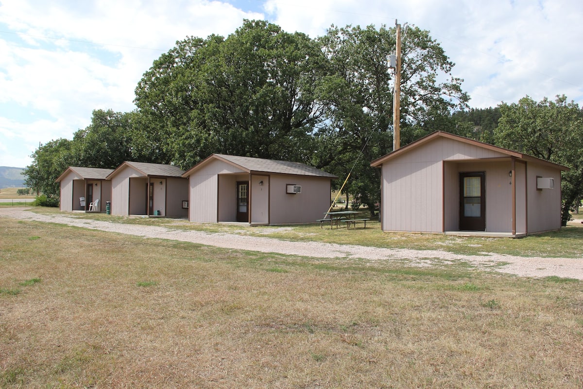 A row of small, single-story cabins with a simple, functional design, each featuring a door and a small window, situated in a grassy area with trees in the background.