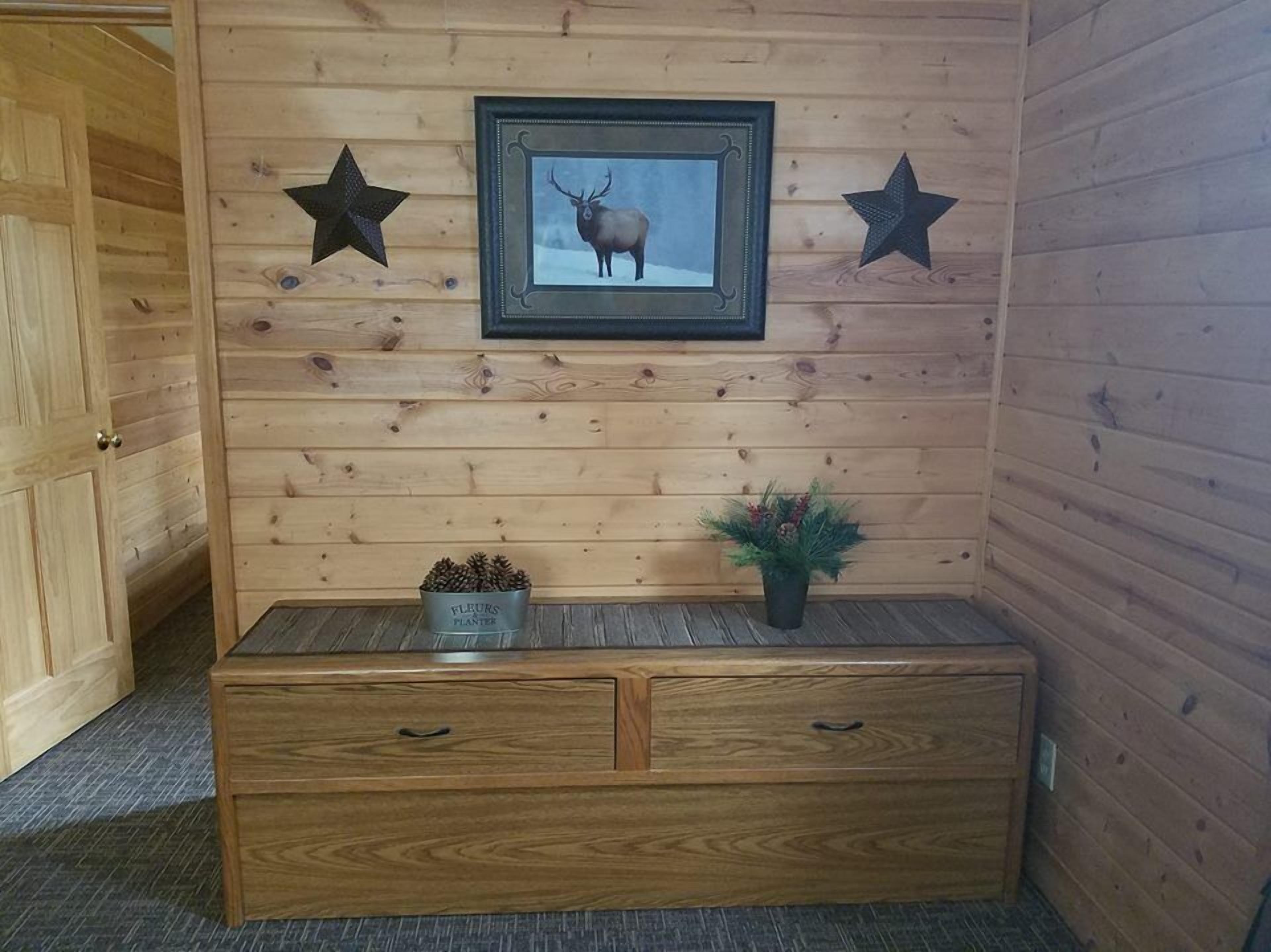 A wooden cabin interior with a framed picture of an elk, two star decorations, a pine cone planter, and a wooden chest of drawers.