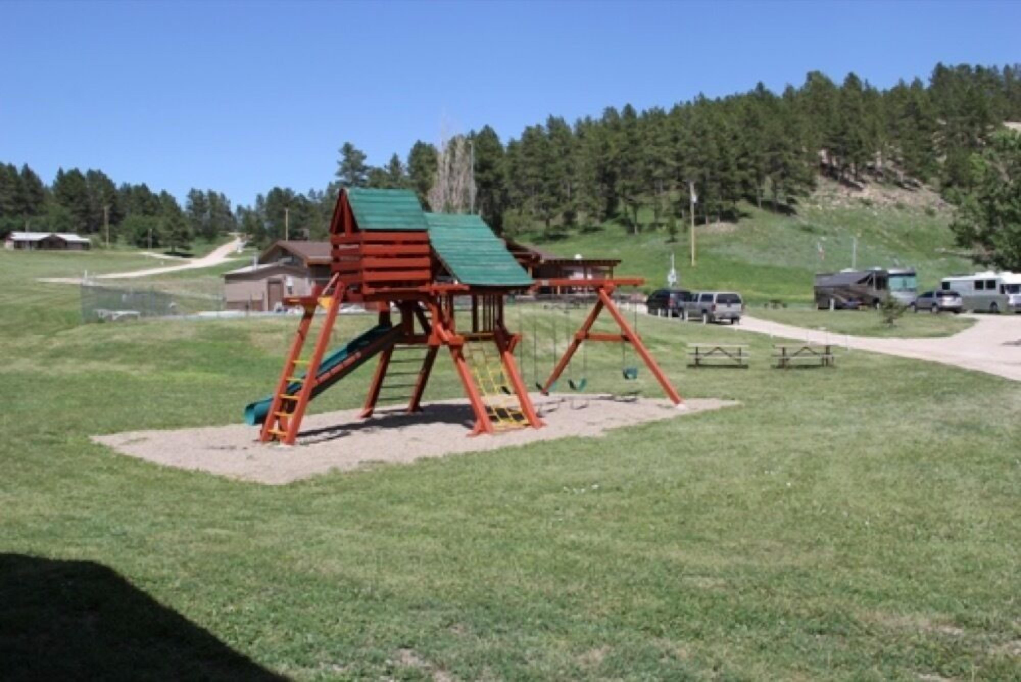 A playground with a slide and climbing structure, surrounded by a grassy field and a forested hill.