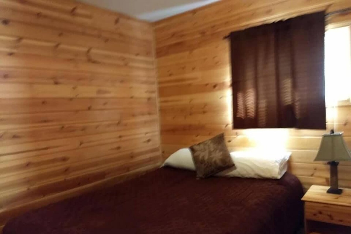 A wooden cabin room with a bed, a nightstand, and a window with curtains.