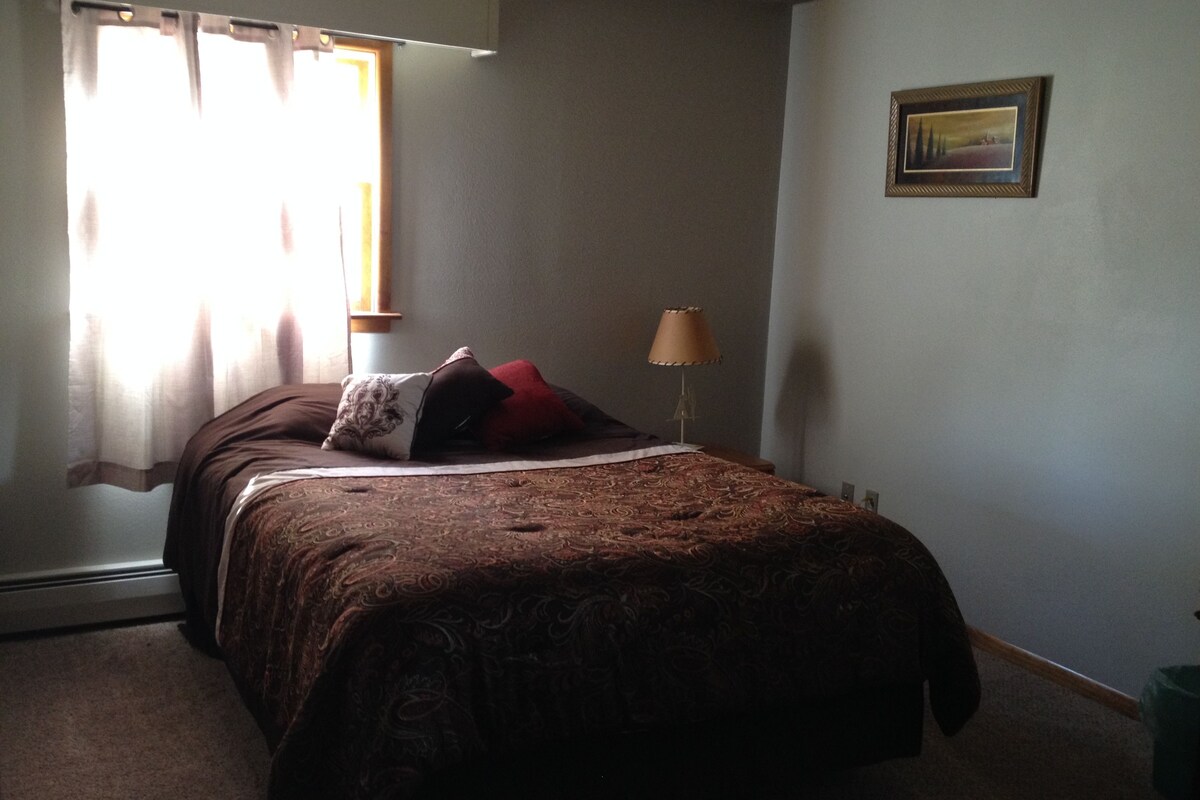 A bedroom with a bed, a lamp, a window with curtains, and a framed picture on the wall.