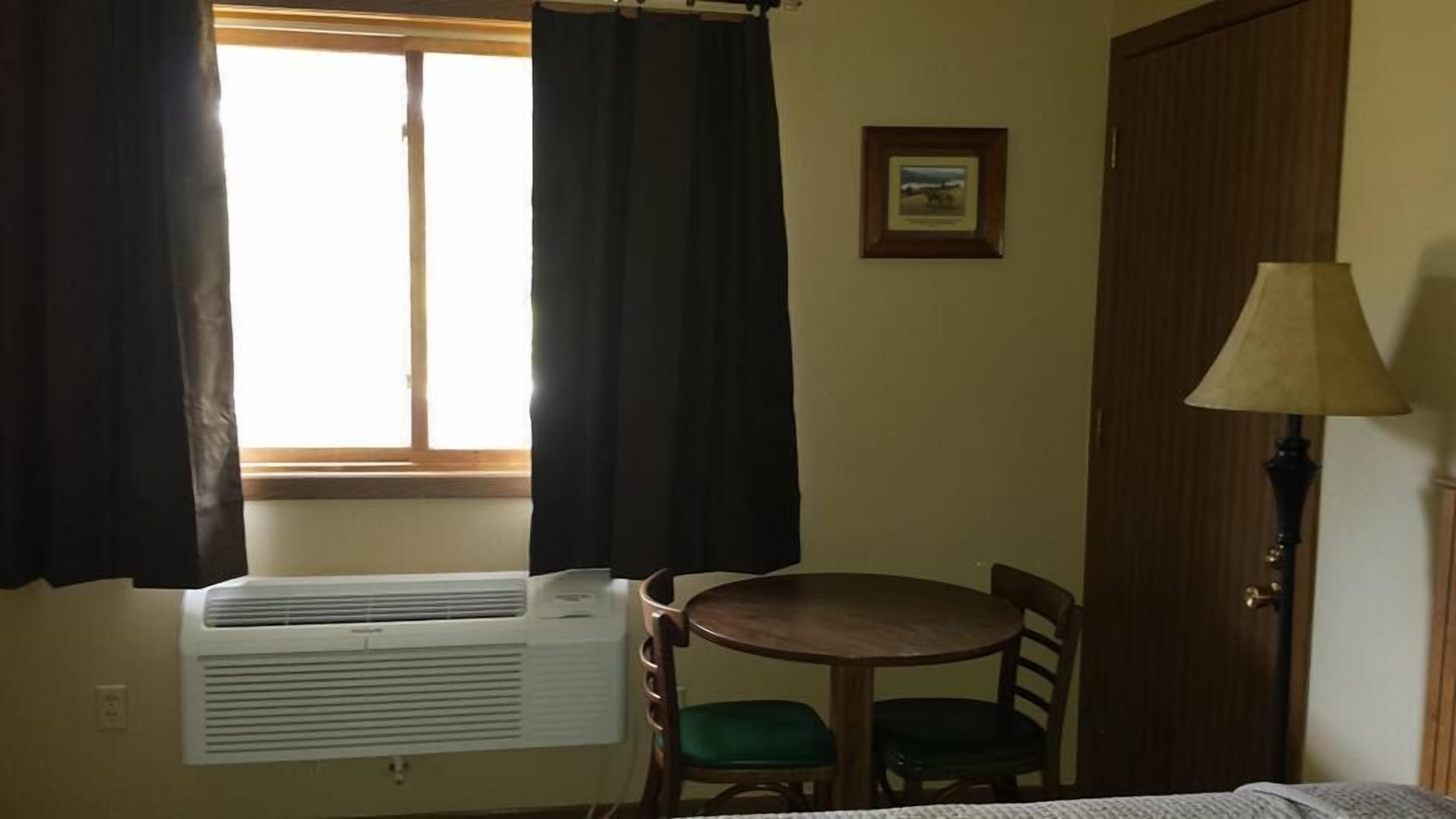 A room with a window, an air conditioner, a table with a chair, a lamp, and a door.