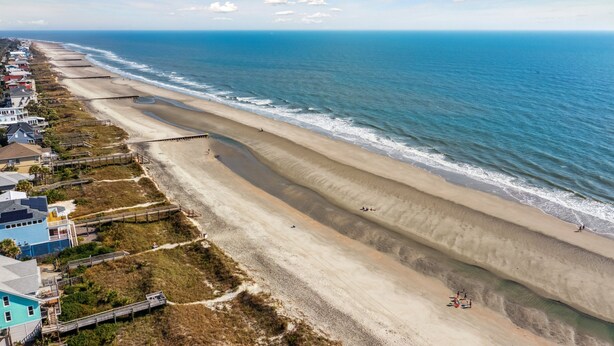 Ein Luftbild eines Strandes mit einer weiten Sandfläche, einigen Gebäuden und einem klaren Himmel.