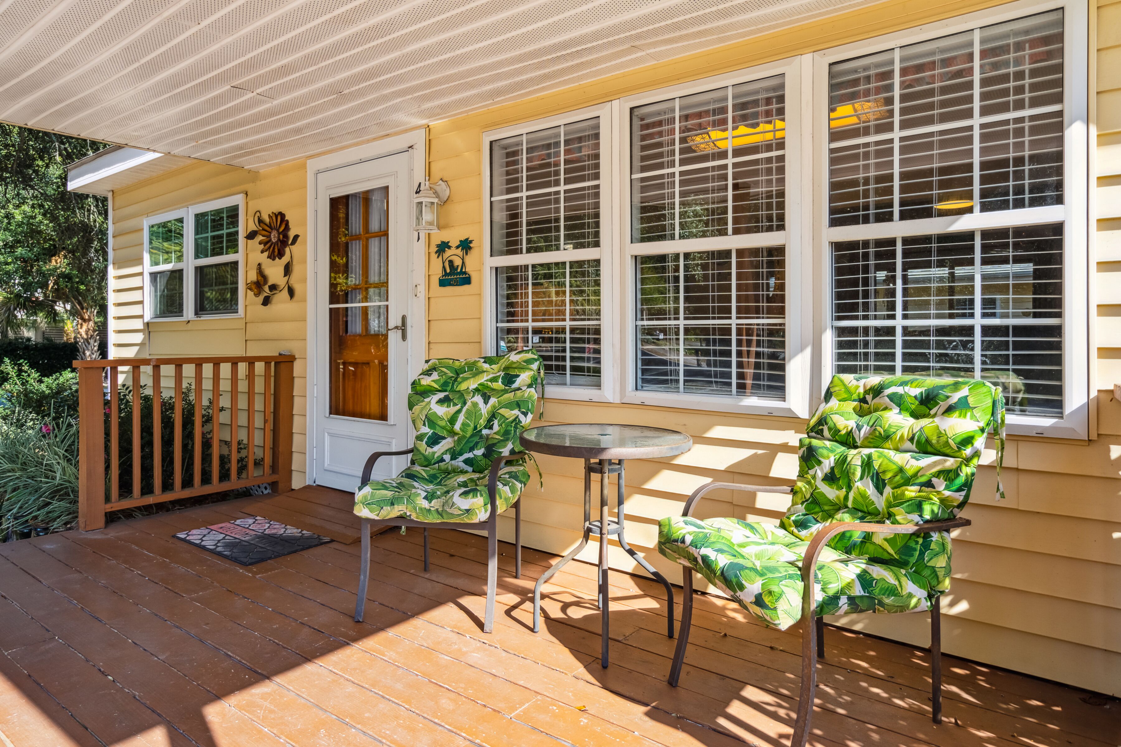 A covered porch with two chairs and a small table, a door with a decorative flower, and large windows with blinds.
