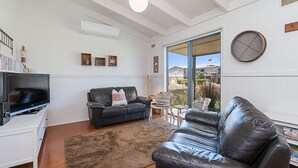 A living room with a leather sofa, a TV, and a large window with a view of houses. - Ocean Spray Retreat - sitting just block from the Southern Ocean (Port Fairy)