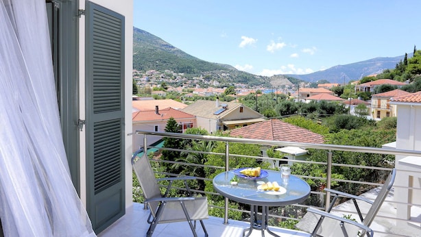 A balcony with a table set for two, overlooking a residential area with red-tiled roofs and green hills in the background. - Ithaca's Poem (Ithaki)
