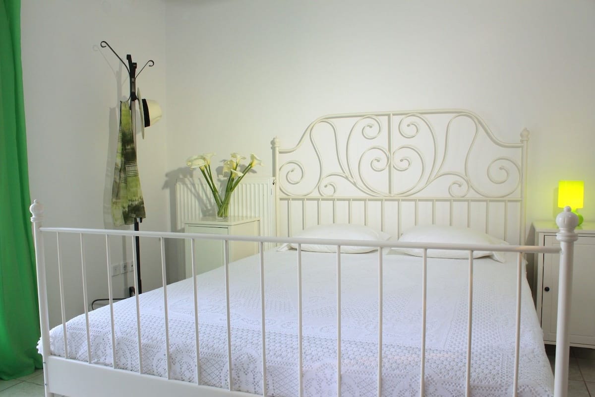 A white metal bed with a decorative headboard, a bedside table with a lamp, and a vase with flowers on a small cabinet.
