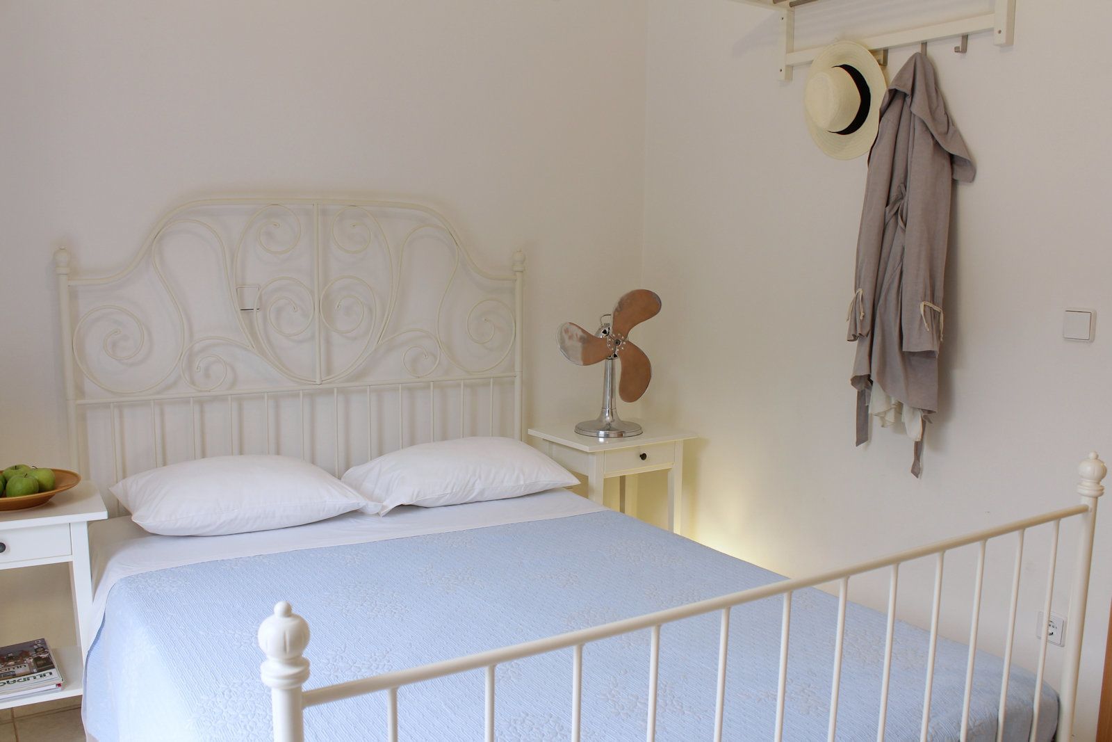 A bedroom with a white bed, a bedside table with a fan, a bowl of fruit, and a coat rack with a hat.