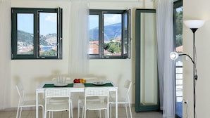 A dining area with a table set for two, two white chairs, a fruit bowl, and a view of a mountainous landscape through large windows. - Ithaca's Poem (Ithaki)