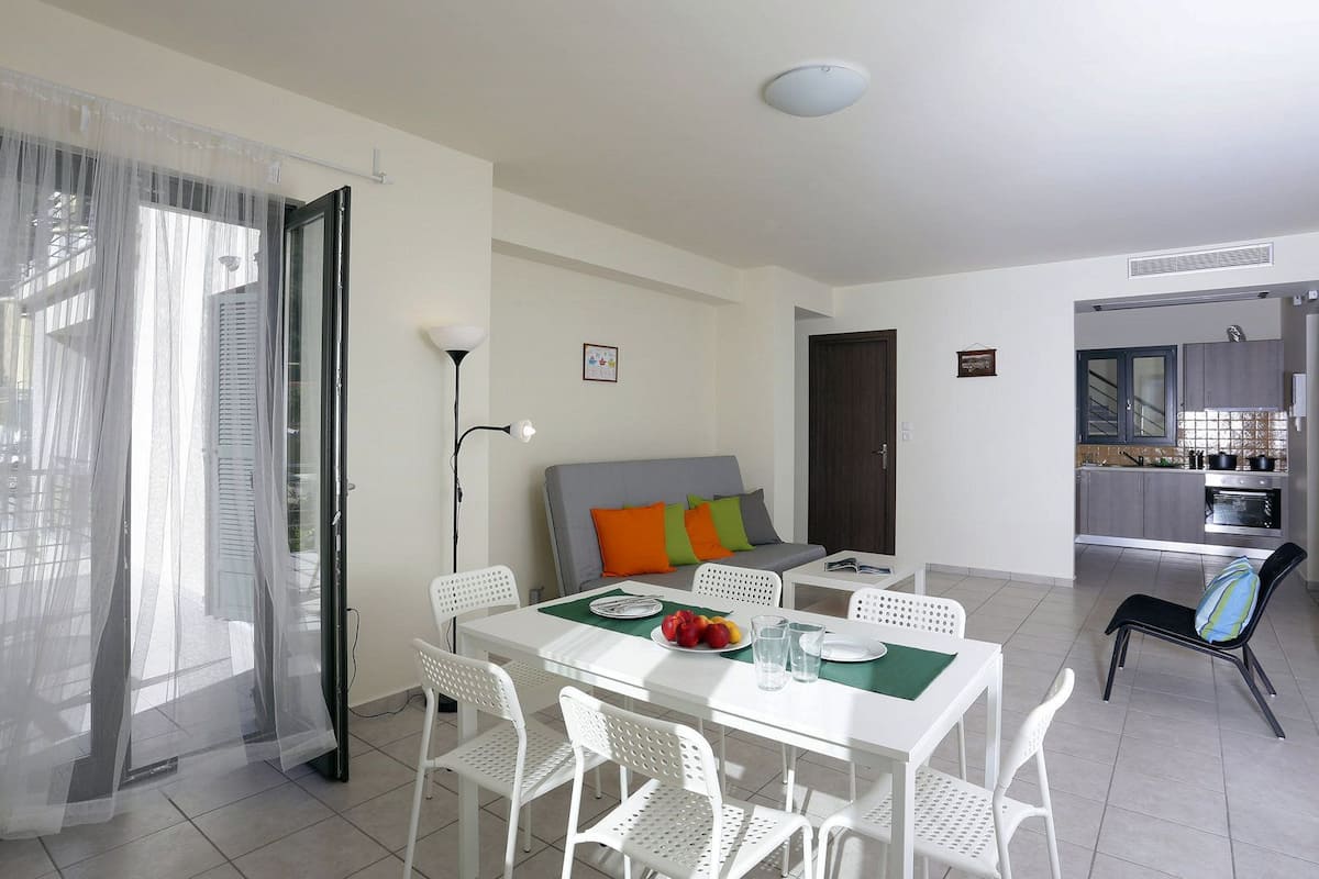 A modern living room with a dining area, a sofa with colorful cushions, a white table with chairs, and a kitchen in the background.