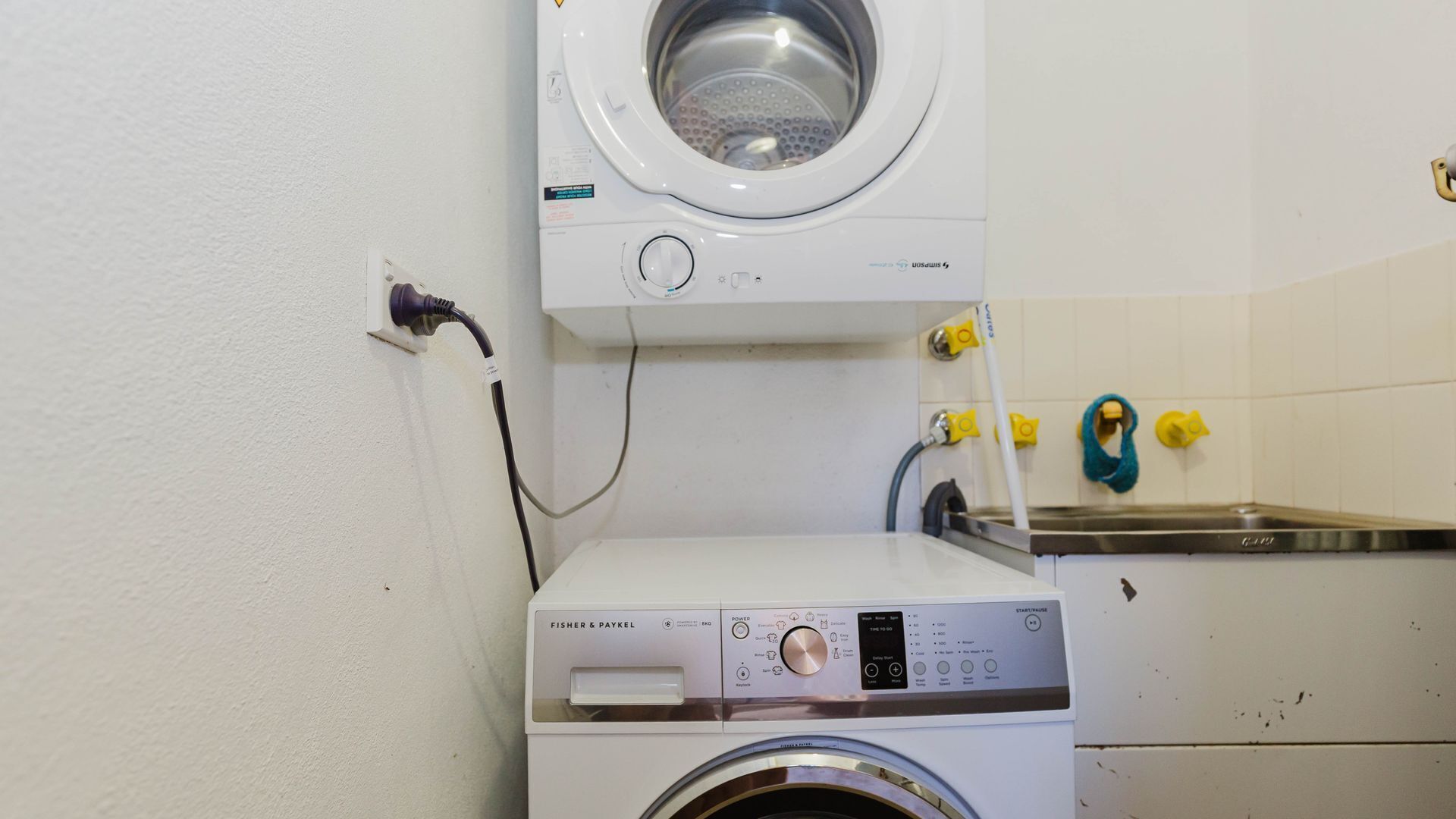 A front-loading washing machine and dryer in a laundry room with a wall-mounted sink and yellow towel hooks.