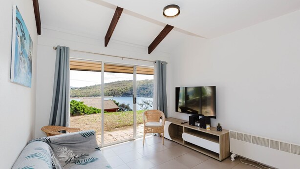A modern living room with a large TV, a sofa, a wicker chair, and a sliding glass door leading to a balcony with a view of a lake and hills. - Kooringa - 1/8 Townsend Street (Jindabyne)